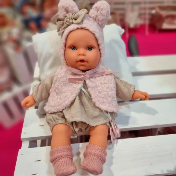 Говореща кукла бебе Кика с меко тяло, с пухкава розова качулка с ушички и панделка, облечена в сива дрешка с розови точки, седнала на бяла поставка