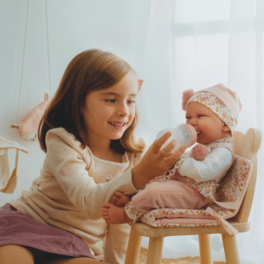 Момиченце храни с шишенце кукла бебе Карла с цветна шапка в дървен стол