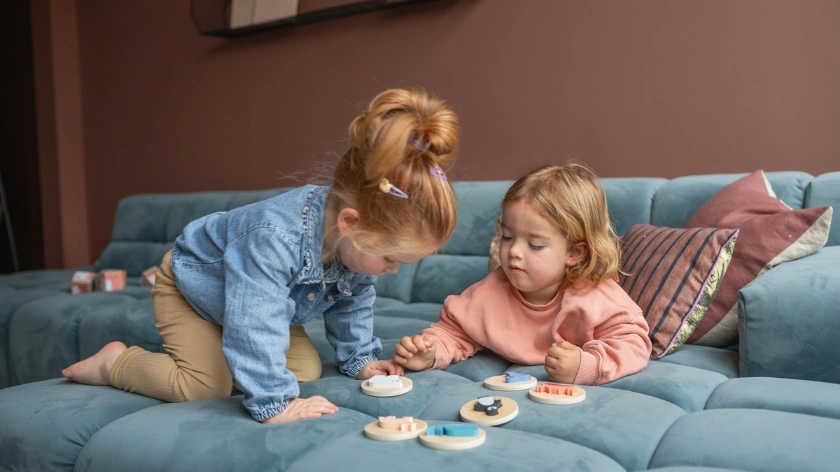 Две малки момичета играят с дървени образователни играчки развиващи фината моторика и паметта на синьо канапе.