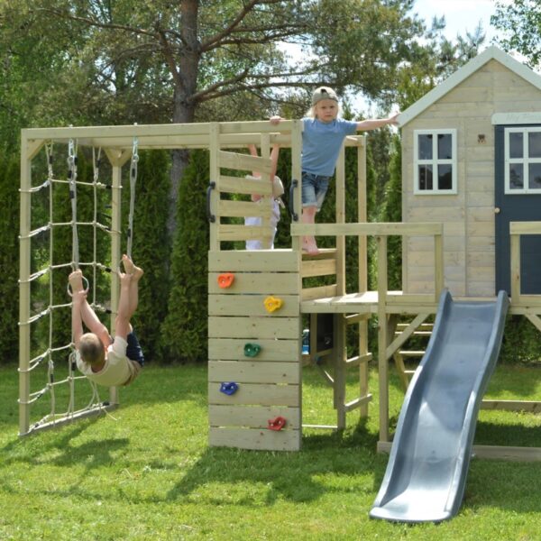 Детски гимнастически модул Wendi Toys -дървен комплекс за игра монтиран върху зелена трева в двора и деца си играят на различните елементи в него едното дете се катери с въже а другото вече е върху стената за катерене