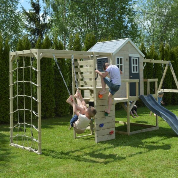 Детски гимнастически модул Wendi Toys -дървен комплекс за игра монтиран върху зелена трева в двора и деца си играят на различните елементи в него едното дете се катери с въже а другото вече е върху стената за катерене