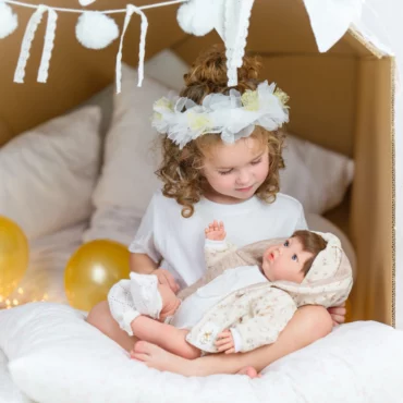 Момиче с цветна диадема държи кукла бебе Marina & Pau, облечена в класически бебешки тоалет от Bellamiestore