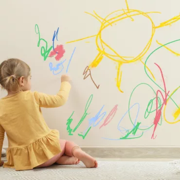 Малко момиче рисува слънце и форми с цветни моливи върху стена, подходящо за ранно детско развитие