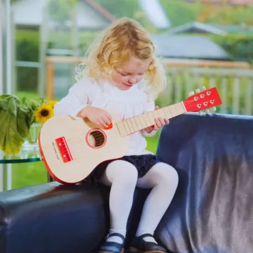 Момиче с дълга руса коса свири на детска дървена китара, червен кант, подходяща за малки музиканти