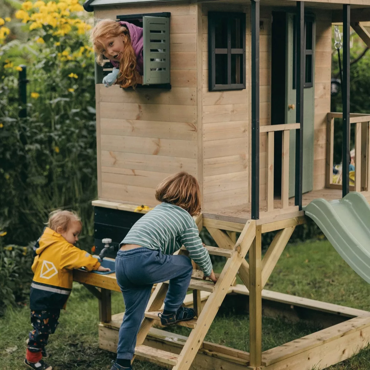 Деца играят на дървена къщичка с пързалка и кухня за забавление на открито